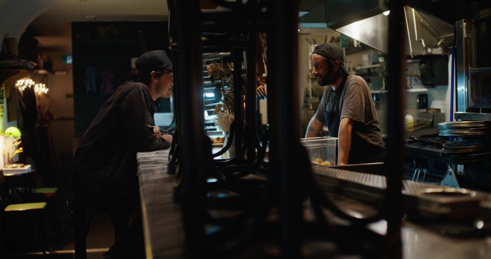 Two actors in a quiet restaurant kitchen after hours, warmed by a pizza oven and practical lamps